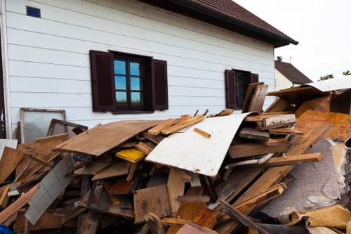 A large pile of assorted construction and renovation debris is situated against the side of a white residential building with dark brown window shutters, which are partially visible. The debris includes wooden planks, panels, and broken fixtures of various lengths and widths, with some pieces showing weathered textures and light to dark brown tones, while others are painted or covered in residual paint. The pile extends across a paved surface, likely part of a driveway or courtyard, with some of the wooden pieces leaning haphazardly and overlapping each other. In the background, the building's exterior wall is smooth with horizontal siding, and part of a sloped roof with red tiles is visible at the top right. The overall scene appears to be a site awaiting rubbish removal, potentially managed by specialists such as Commercial Waste Removal Millbank, specialists in waste clearance in the area near Millbank or the greater Westminster region, with natural daylight illuminating the scene and highlighting the textures and colors of the debris.