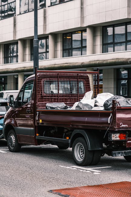 The image shows the rear side of a white commercial van parked on a street near a building with large glass windows and a dark-colored wall. Attached to the back of the van is a metal ladder rack, and