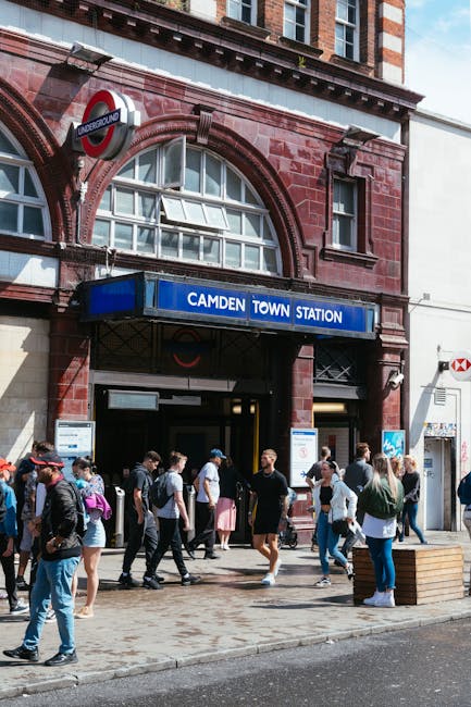 Photograph of the entrance to Camden Town Underground Station, built with red brick and featuring large arched windows. The station sign is prominently displayed in blue with white lettering, reading 