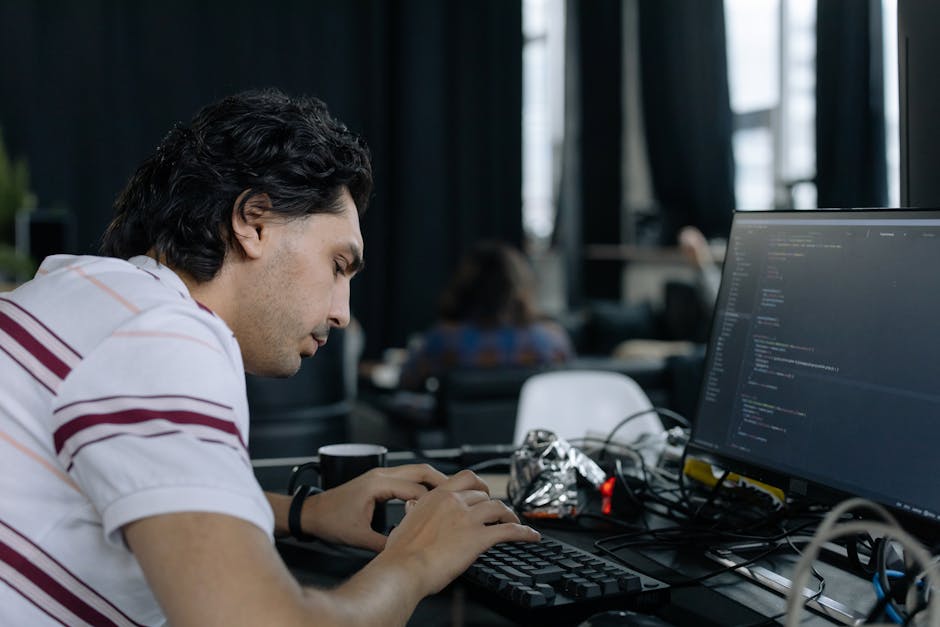 A young man with dark, curly hair and wearing a white shirt with red and purple horizontal stripes is seated at a desk in an indoor office environment. The desk is cluttered with various items, includ